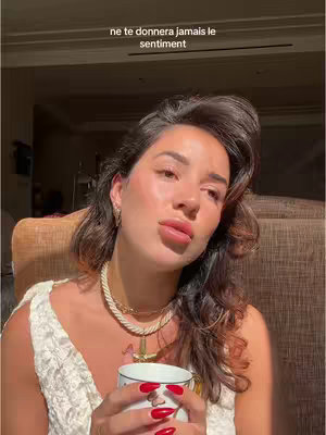 A woman with light skin and dark, wavy hair is looking at the camera. She is holding a white mug with a floral design and has red nail polish. The image is warm-toned, with a focus on the woman's face and upper body. The background is slightly blurred, with warm lighting.
