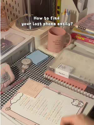 A flat lay of a desk setup with various items. In the center is a pink mug with text. A notebook with a pink cover, pens, sticky notes, a small jar with items are present, layered with other items. The background looks like a desk with some books on the side.