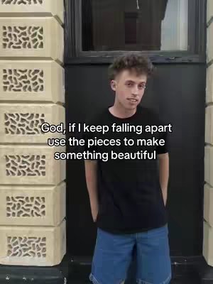 A young man stands in front of a building's exterior. Above him, text overlays read, 'God, if I keep falling apart use the pieces to make something beautiful.' The man wears a black t-shirt and jean shorts, with hands in his pockets. The background is mostly black with vertical stone details, and parts of the image are out of focus.
