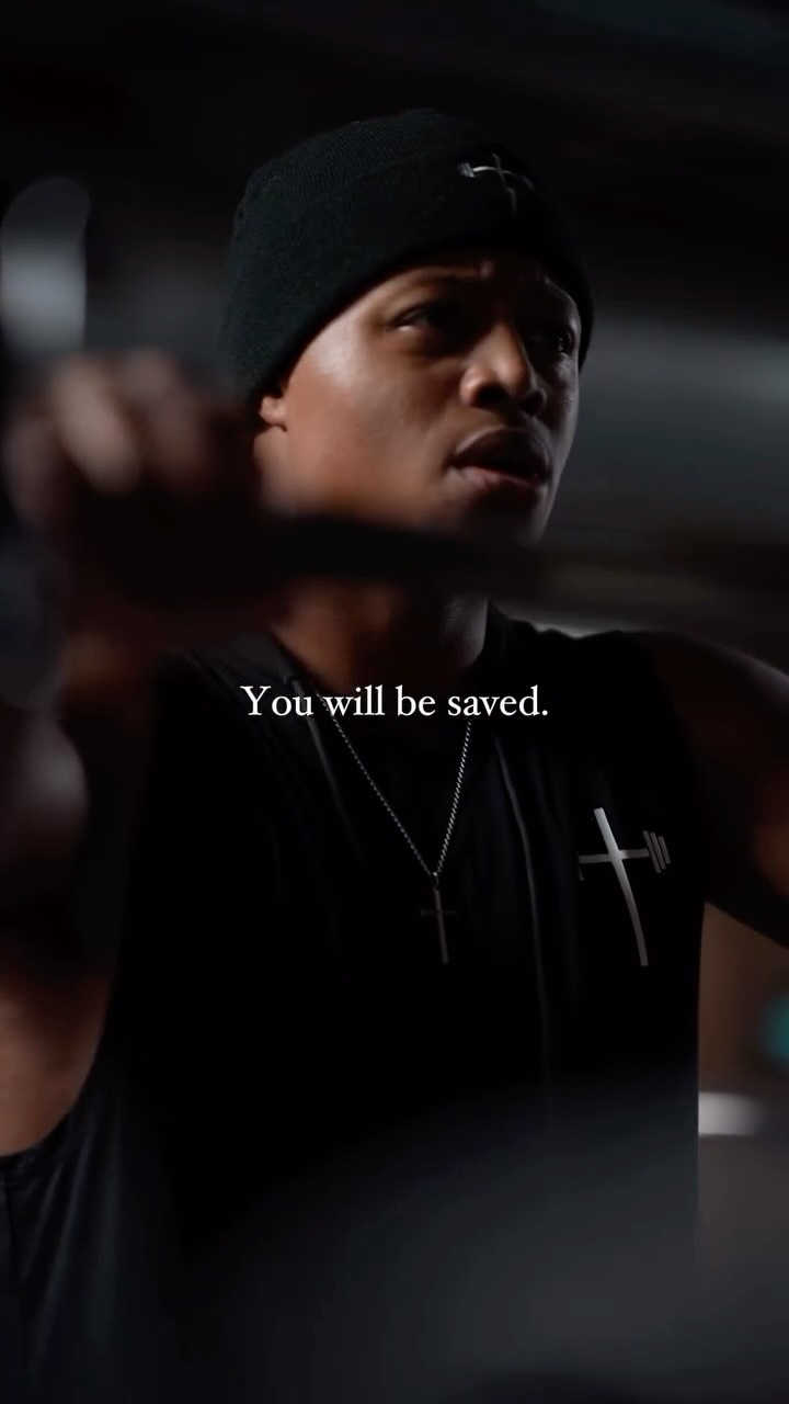 A close up shot of a black man wearing a black beanie and a silver cross necklace, lifting a weight. The image is dark, with the focus on the man's determined expression. Text overlayed onto the image reads "You will be saved"
