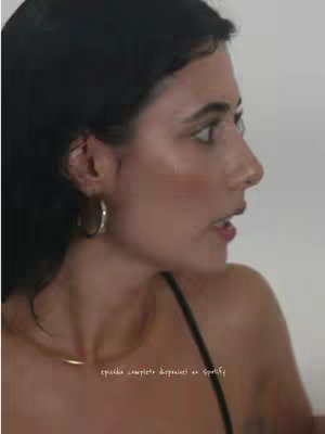 A side profile shot of a woman looking to the right. She has dark hair, a silver hoop earring, and is wearing a black spaghetti strap top. She has a light neutral background.