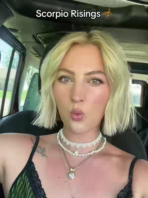 A selfie-style shot of a woman in a car. She has blonde hair and is wearing a black and green top, with multiple necklaces. The background indicates she is in the driver's seat, surrounded by the car's interior.