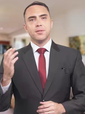A man in a black suit with a red tie is speaking, his right hand gesturing. The background is slightly blurred, suggesting an interior setting.