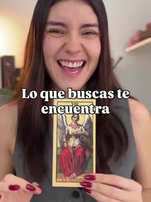 A woman with long brown hair smiles broadly, holding a tarot card that features an angelic figure. The background is softly lit with warm tones. Overlaid text reads: 'Lo que buscas te encuentra'.