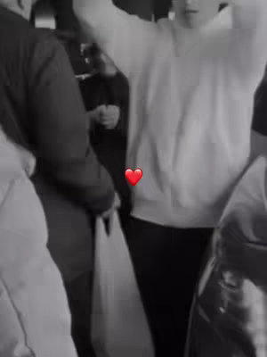 A black and white photo with a heart emoji. The scene captures multiple people in a crowded place, with a person in a white hoodie and dark pants as the central focus. The color palette is limited to shades of gray, enhancing the simplicity of the image.