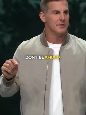 A man in a tan bomber jacket and white shirt is the focal point, delivering a message. The background appears to be blurred, suggesting a large room or stage.