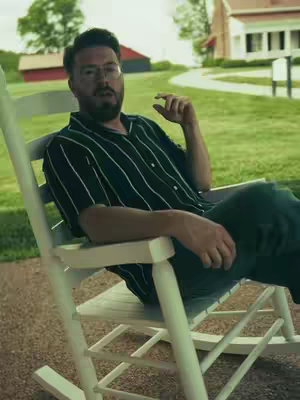 A man with a beard and glasses sits in a white rocking chair on a porch. He is wearing a black and white striped shirt and green pants. The background shows a green lawn, a red barn, and a white house with a red roof.