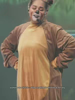 A person in a brown and tan bear-like costume, with closed eyes; face paint and a microphone; set against a blurry, green-toned background.