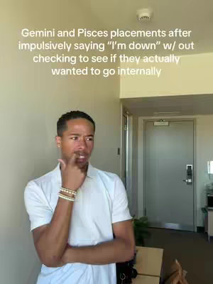 A man with short, dark hair and a light complexion is shown from the chest up. He is wearing a white collared shirt and a few bracelets. He is standing in front of a neutral wall and door, with a small plant and a table visible in the background, and he is looking to his right and touching his index finger to his lips in a thoughtful gesture.