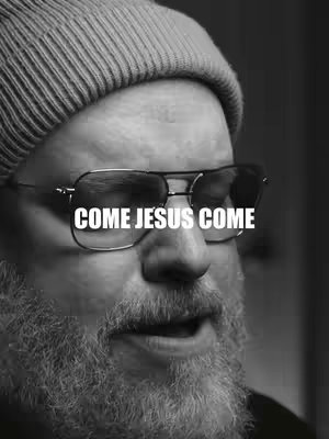 A black and white close-up shot of a man wearing a beanie and glasses. The glasses have a clear lens highlighting the text overlay of \"COME JESUS COME\" in a white bold font. The lighting is even, illuminating the man's face, beard, and glasses. The background is dark, without details.
