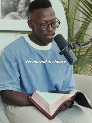 A man wearing glasses and a blue shirt sits on a couch, holding an open book with red edges and a finger marking his place while speaking into a microphone. In the background is a plant and a black and white photo.