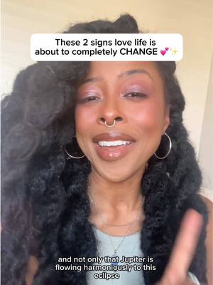 A cover image featuring a dark-skinned woman with long, curly black hair. She is smiling and looking upwards with a friendly expression. Her face is the central focus, with a partially visible finger pointing upwards. She wears a nose ring and hoop earrings. She is wearing a light top to enhance visibility. An overlaid text in a white rectangular bubble states that two signs of the zodiac are about to experience love-life changes. The background is a light-colored surface.