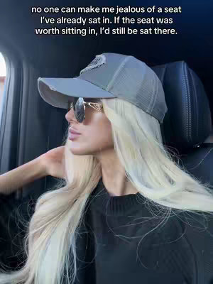 A woman in a car, wearing sunglasses and a baseball cap, looking to the side. The lighting suggests it's daytime. Text overlay is at the top.