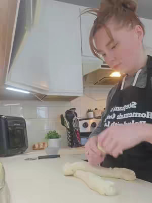 A woman in a kitchen is shown in a medium shot. She has red hair in a bun and is focused on working with dough on a countertop. The background includes kitchen cabinets, a microwave, and a toaster.