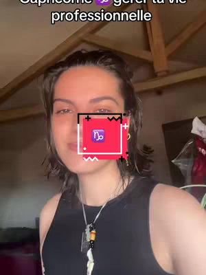 A medium shot of a young woman looking at the camera, with text above her head and a zodiac symbol over her face. She has dark hair and is wearing a black top and a necklace. The background appears to be indoors.