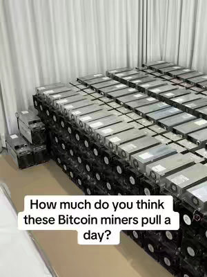 A stack of server-like machines, potentially for cryptocurrency mining, fills the frame. The machines are rectangular with fans visible on the front. The background is a simple white curtain.