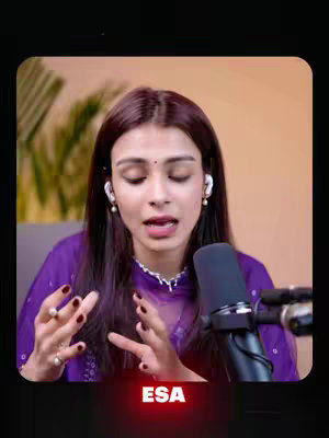 A woman with long dark hair, wearing earphones and a purple shirt, is speaking into a black microphone. Her hands are gesturing. Behind her is a blurred green background.