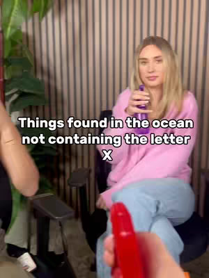 A young woman in a pink sweatshirt, holding a purple spray bottle and sitting in a chair, looks towards the camera. The background is blurred, with a vertical line pattern. The person in the foreground is holding a red bottle and the view is from their perspective. Text overlay reads 'Things found in the ocean not containing the letter X' in white letters.