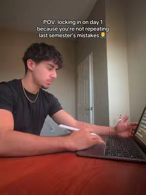A young man studies at a desk with a laptop open in front of him. He's wearing a black shirt and a gold chain. The scene is lit by natural light.