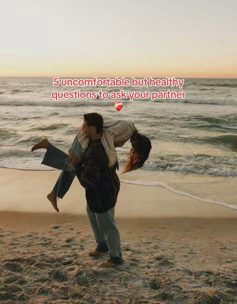 A man carries a woman on his shoulders at the beach. The man is wearing a dark jacket and grey pants, while the woman is wearing a white top and jeans. They are standing on the sand next to the water. The sky is a gradient of orange and yellow, suggesting a sunset.