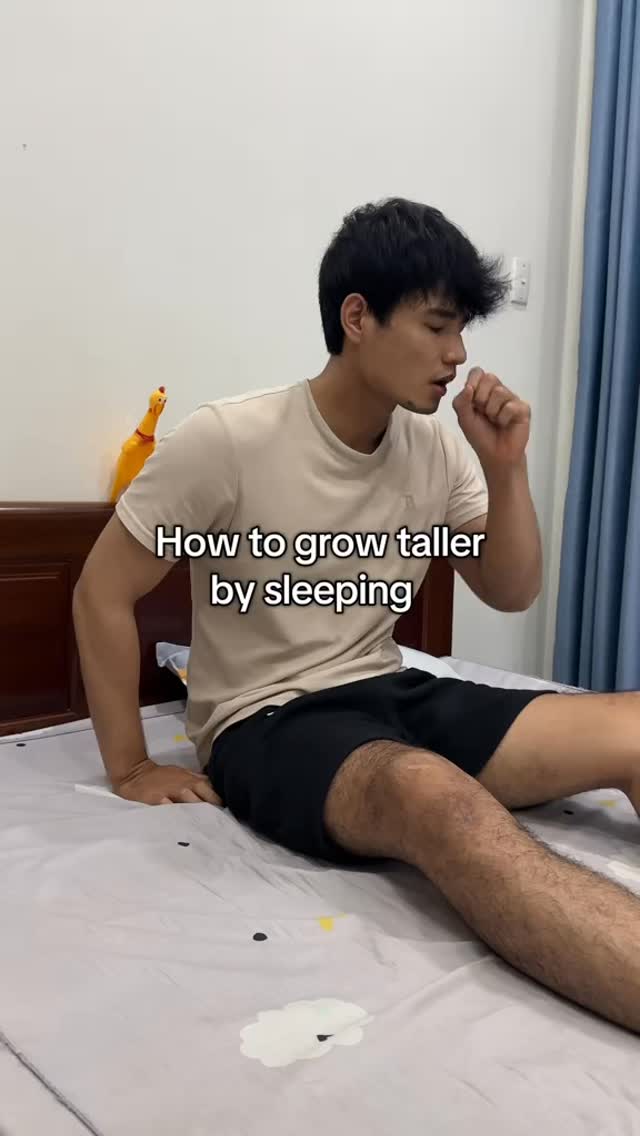 A young man is sitting on a bed with a light gray patterned bedsheet. The man is wearing a light beige t-shirt and dark shorts. Text overlayed on the image reads, 'How to grow taller by sleeping'. He is sitting up and appears to be gesturing with his right hand near his face, possibly in a thoughtful pose. Behind him on the wall is a yellow toy.