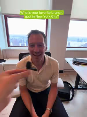 A man in a neutral-colored shirt is seated in a chair while a person’s hand, holding a microphone, is visible in the lower left corner. Overlaid on the image is a textual question in a green banner that is asking, "What's your favorite brunch spot in New York City?"