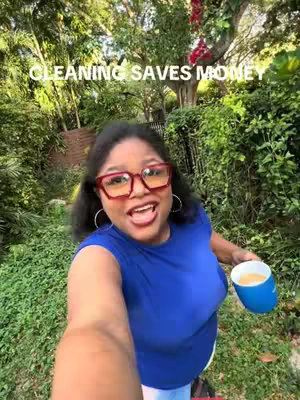 A woman with dark hair and glasses smiling at the camera while holding a blue cup outdoors. The background includes lush greenery and a tree.