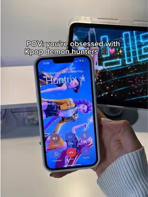 A hand holding a phone with the lock screen showing a colorful anime-style wallpaper. In the background, there is a laptop with a neon design and a blurred background.