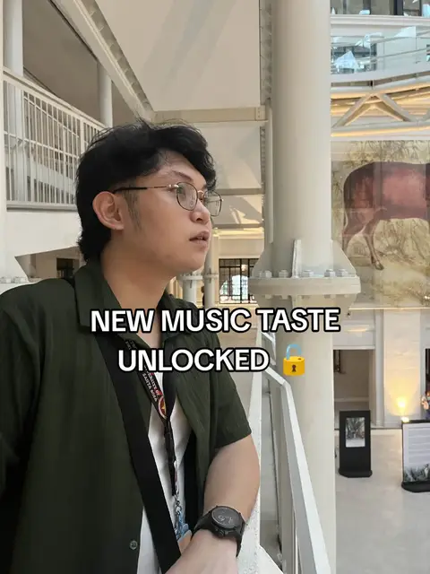 A young man in casual attire stands on a balcony overlooking a bright, well-lit hall. He looks thoughtful to the side. Text overlays read 'NEW MUSIC TASTE UNLOCKED' with a lock and key emoji over a textured background.