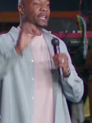 A man wearing a light blue button-down over a pink t-shirt speaks into a microphone.