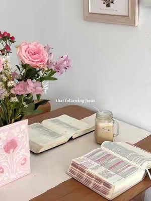 A photo presents a serene interior scene, likely a home setting, with a wooden table serving as the focal point.  On the table, an opened Bible, a second book, and a clear glass jar with what appears to be a beverage and a straw are visible, suggesting a moment of quiet contemplation or study. A bouquet of pink and purple flowers enhances the setting. The background is somewhat blurred, featuring a framed picture and a white wall, adding to the photo's soft, inviting aesthetic.