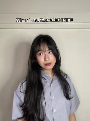 A young woman with long black hair and bangs stands in front of a plain white wall. She is wearing a light blue button-down shirt. Text overlay reads: "When I saw that exam paper".