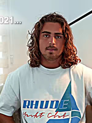 The image features a young man with wavy brown hair, wearing a white t-shirt with the text "RHUDE Yacht Club" in bold blue and red letters. He is looking directly at the camera with a neutral expression. The background is plain, suggesting a neutral setting.