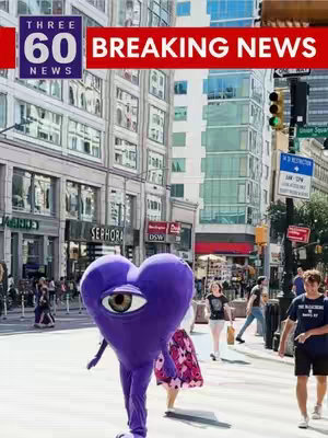 A street scene featuring a person in a heart-shaped mascot costume with a large eye on it, walking towards the viewer. The background includes city buildings, traffic, and pedestrians. Text overlays read "THREE 60 NEWS" and "BREAKING NEWS".