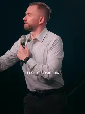 A man with short hair, a beard, and a light-colored shirt is speaking into a microphone. The lower left corner of the image shows text indicating that it tells us something. The background is a dark gradient.