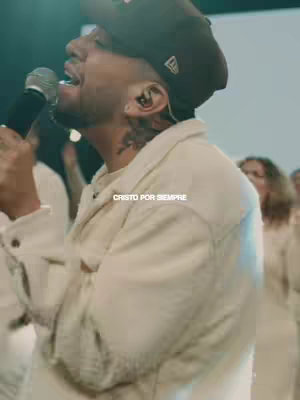 A man with a microphone singing in front of a blurred background. He wears a brown cap and a white shirt, and a tattoo can be seen on his neck. The text "CRISTO POR SIEMPRE" is overlayed.