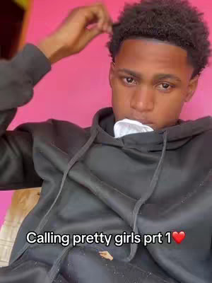 A young man with short, dark, curly hair is shown in a selfie-style shot. He's wearing a black hoodie and appears to be holding a piece of cloth near his mouth. A vibrant pink wall serves as the background. Text overlay reads: Calling pretty girls prt 1 ❤️.