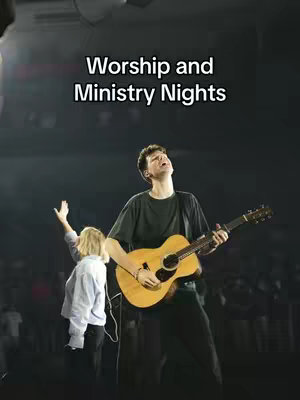 Image is of a stage performance with two people. The main focus is on a man playing an acoustic guitar, with light shining down from above. Behind him, another person stands with their arms raised. The background is dark with stage lighting.