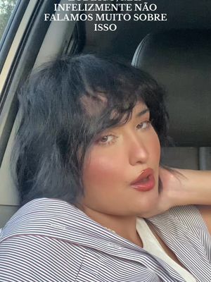 A selfie of a person sitting in a car. The person is fair-skinned with short, dark, wavy hair and full lips. There is text overlaid on the top that's partially out of frame. The person is wearing a pinstriped blouse and looks to the side with a neutral expression.