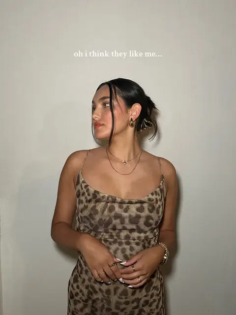 A young woman with dark hair styled in a half-updo is shown looking over her shoulder. She wears a leopard print dress with thin straps, gold jewelry, and multiple rings. The background is a plain, light-colored wall. Text overlay reads 'oh i think they like me...'