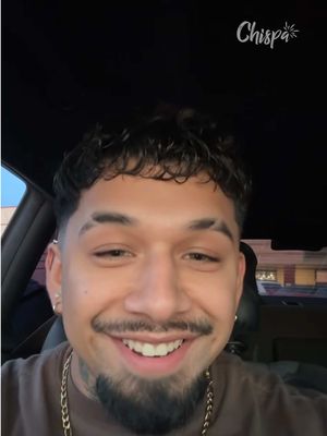 A selfie captured from a car's front seat shows a man smiling at the camera. He has neatly styled, short, dark hair and a short beard. He is also wearing a gold chain around his neck. The background is blurred, suggesting he is driving. The image has a warm tone, enhanced by natural light.
