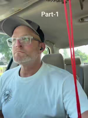 A man in a car, wearing glasses, a hat, and a light-colored shirt. The car interior is visible with red straps hanging down and a sign labeled 'Part-1' in the upper right corner.