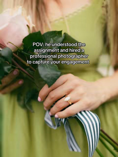 A close-up shot of a person's hand holding a bouquet of pink roses, showcasing an engagement ring prominently displayed. The hand is in the foreground, with a blurred background featuring a green dress and foliage. Text overlay on the image reads 'POV: he understood the assignment and hired a professional photographer to capture your engagement'.