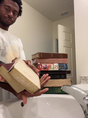 A man in a white shirt is holding and looking at several books near a bathroom sink. A stack of books sits next to him. The lighting is neutral.