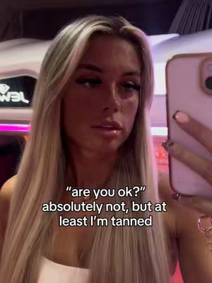 A selfie-style shot featuring a blonde woman in a tanning bed. She's holding a phone to record and appears to be slightly upset. The background shows the interior of the tanning salon with visible equipment and neon lighting that casts a pink hue on the surroundings. Text overlayed reveals the internal monologue of the creator.