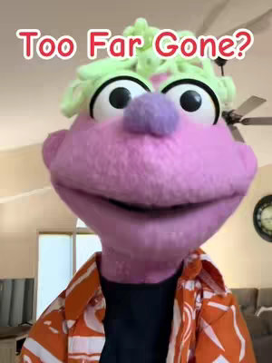 A close-up shot of a puppet with large eyes and colorful features. The puppet has a pink face, a purple nose, and green, curly hair. It is wearing an orange and white patterned shirt. The background is blurred, showing a ceiling fan and room interior.