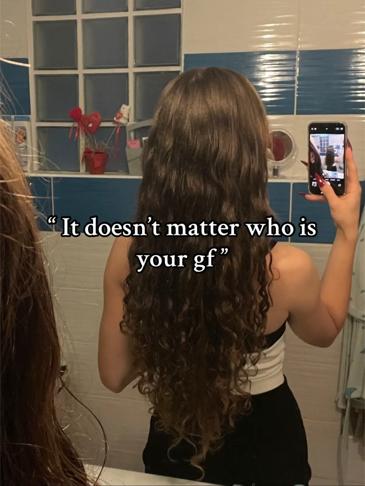 The image shows a woman from the back, holding a phone in front of a bathroom mirror and taking a selfie. She is wearing a white top and black attire. Her long, curly brown hair is prominent. In the mirror's reflection, a decorated wall is visible. Text overlayed above the mirror states "It doesn't matter who is your gf".