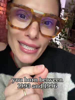 A close-up shot of a woman wearing large, patterned eyeglasses, her expression is a subtle smile as she looks directly at the camera. The bottom of her face reveals her lips and teeth, and the text overlay reads, "you born between 1993 and 1996". The background is softly blurred with suggestions of other objects or decor.
