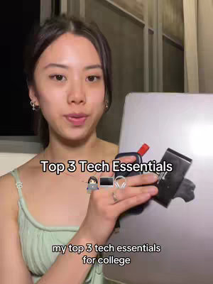 A young woman with dark hair, wearing a light green top, is centered in a selfie-style shot. She is holding a silver laptop with a black item on top, and appears to be presenting the laptop and accessories to the viewer. Beside her, the backdrop is dim and partially out of focus, adding depth. The overall lighting is moderate with a slight emphasis on the subject.