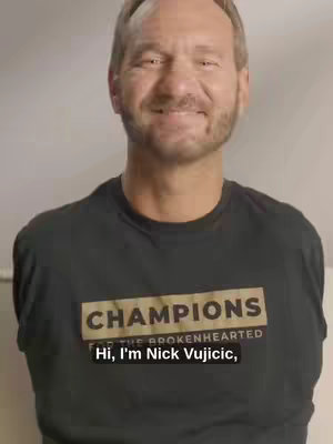 A medium shot of a man smiling at the camera. He has a short beard and gray hair. He is wearing a black t-shirt with the word CHAMPIONS in large tan letters and the words FOR THE BROKENHEARTED beneath it. Below the man is a text overlay in a bold white font that says, "Hi, I'm Nick Vujicic," and is enclosed in a black rectangle.